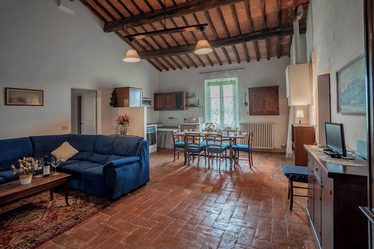 A spacious living room with a blue sofa, a coffee table with flowers, a dining area with wooden chairs, and a kitchen in the background.
