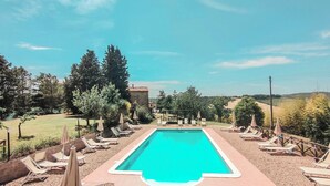 A rectangular swimming pool with a clear blue water, surrounded by lounge chairs and umbrellas, set against a backdrop of trees and a stone building. - Agriturismo La Ripolina (Buonconvento)