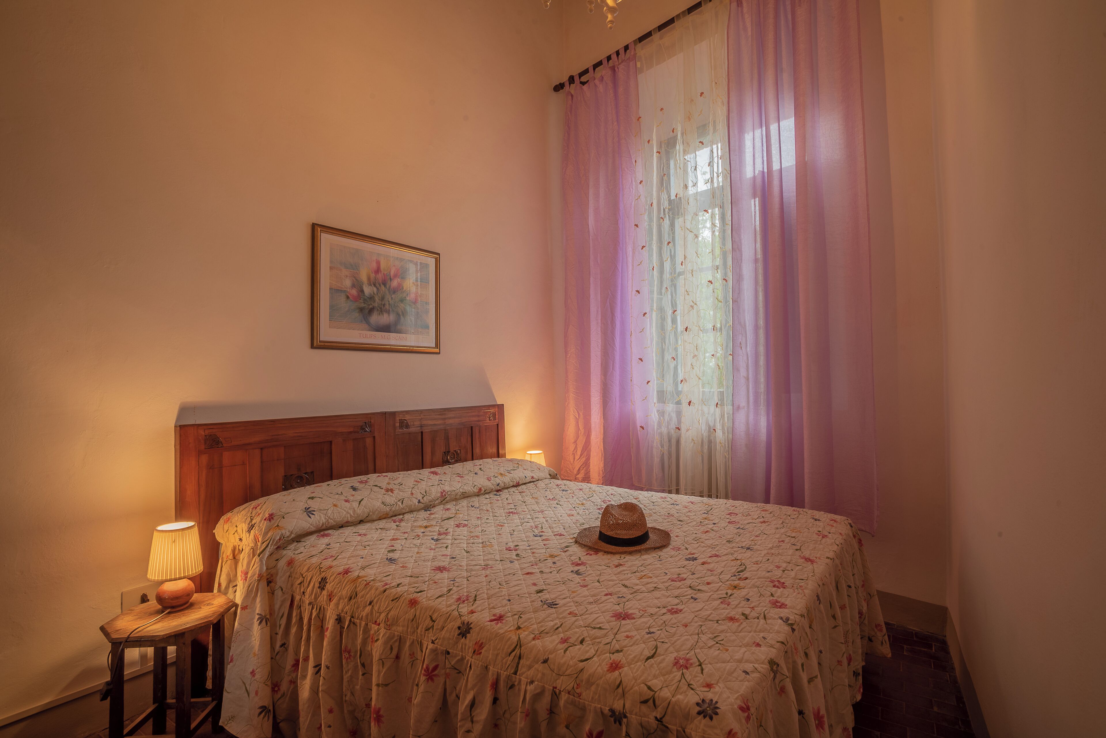 Un dormitorio con cama, mesita de noche, lámpara, sombrero, cuadro en la pared y ventana con cortinas.