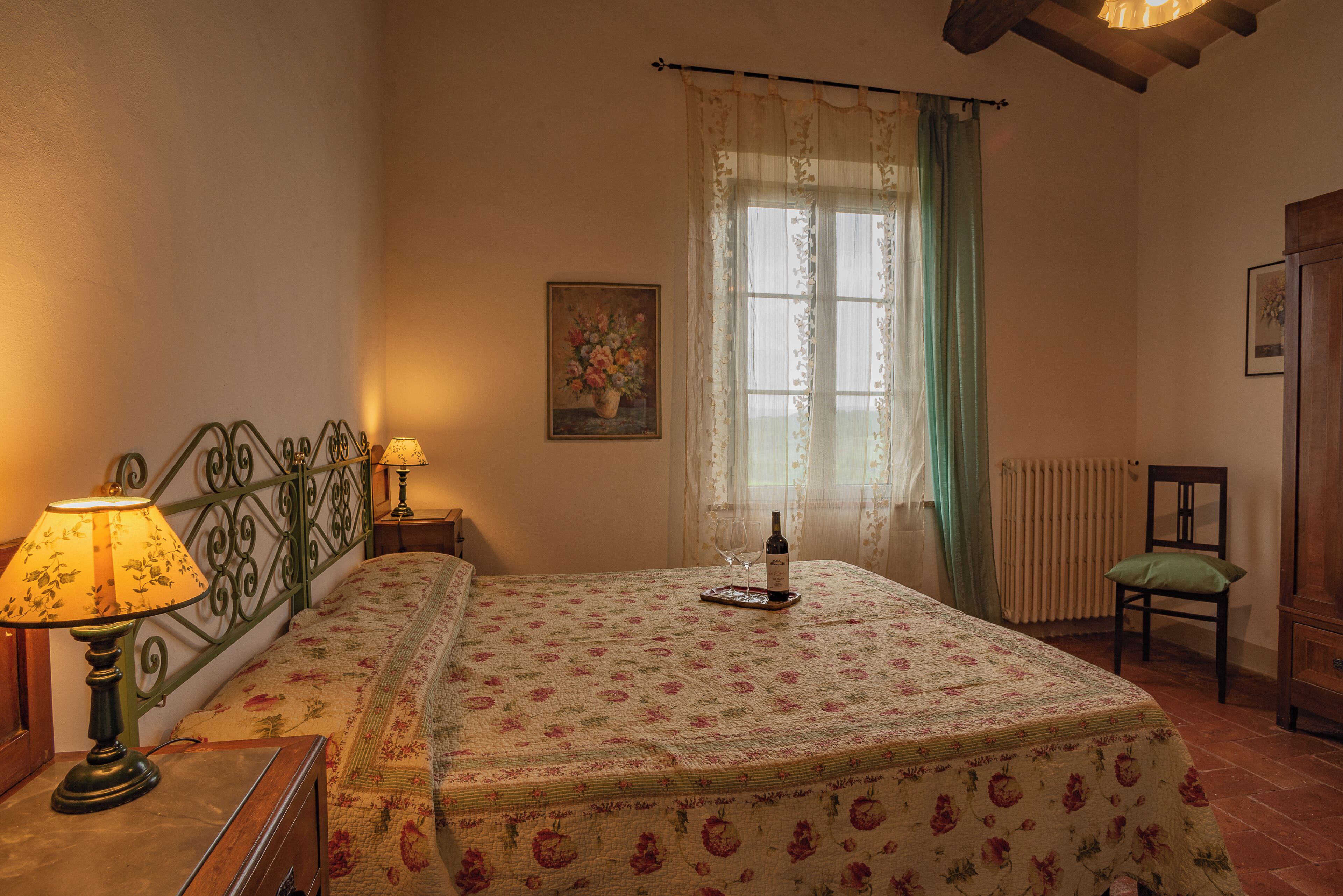 Un dormitorio con colcha estampada, una mesita de noche con lámpara, una ventana con cortinas y una silla.