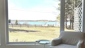 A room with a large window offering a view of a body of water and a dock. There is a striped armchair and a small table with a tray. - Waterfront 2 BR cottage on 1 acre private lot walking distance to beaches! (Stanhope)