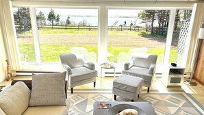 A living room with a sofa, two armchairs, a coffee table with a bowl of snacks, and a view of a lake with a wooden deck and cyclists. - Waterfront 2 BR cottage on 1 acre private lot walking distance to beaches! (Stanhope)