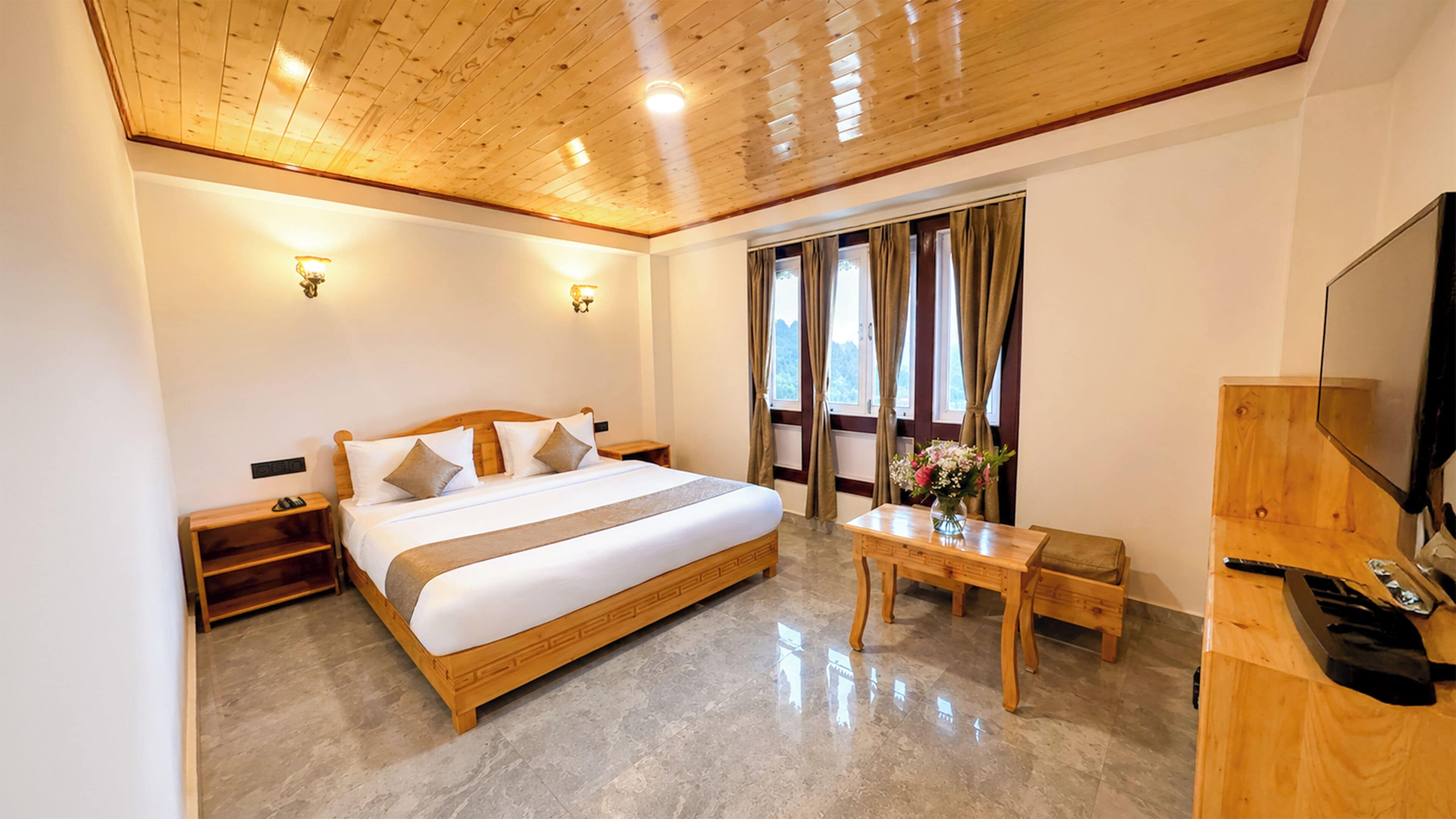 A hotel room with a wooden ceiling, a large bed, a bedside table, a TV, and a window with curtains.