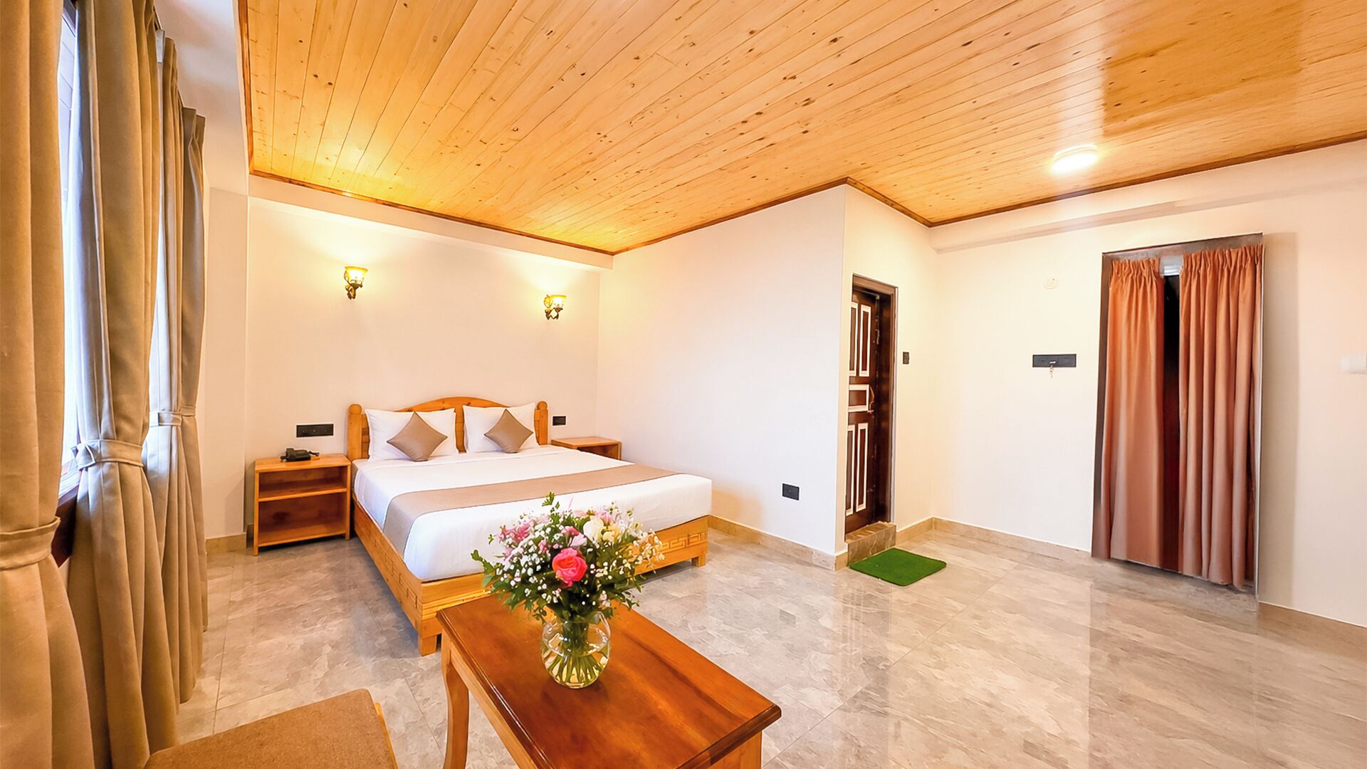 A hotel room with a wooden ceiling, a bed with white linens, a bedside table, a nightstand, a green mat, and a wooden bench.