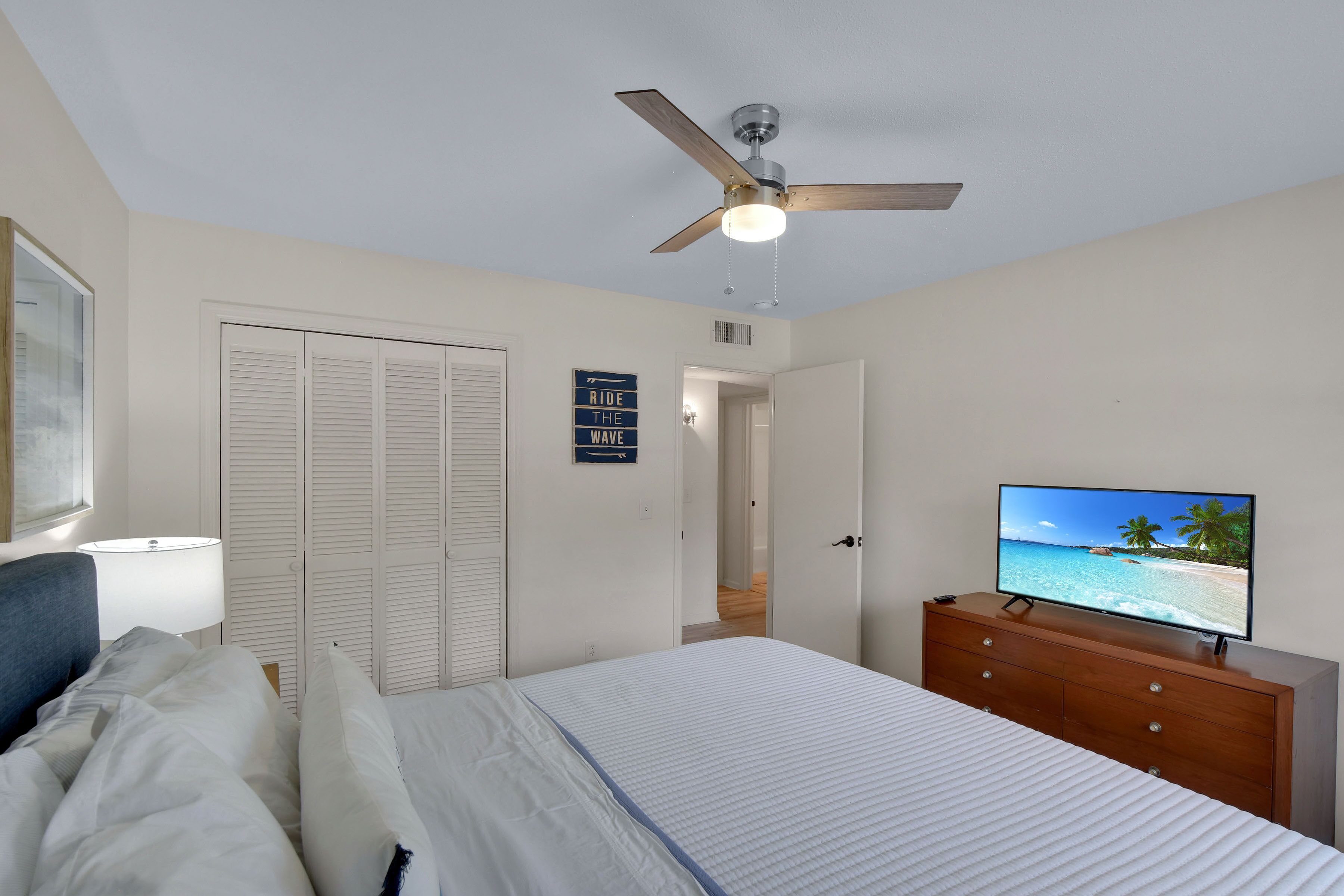 A bedroom with a bed, a dresser, a television displaying a beach scene, a ceiling fan, and a door with a sign.
