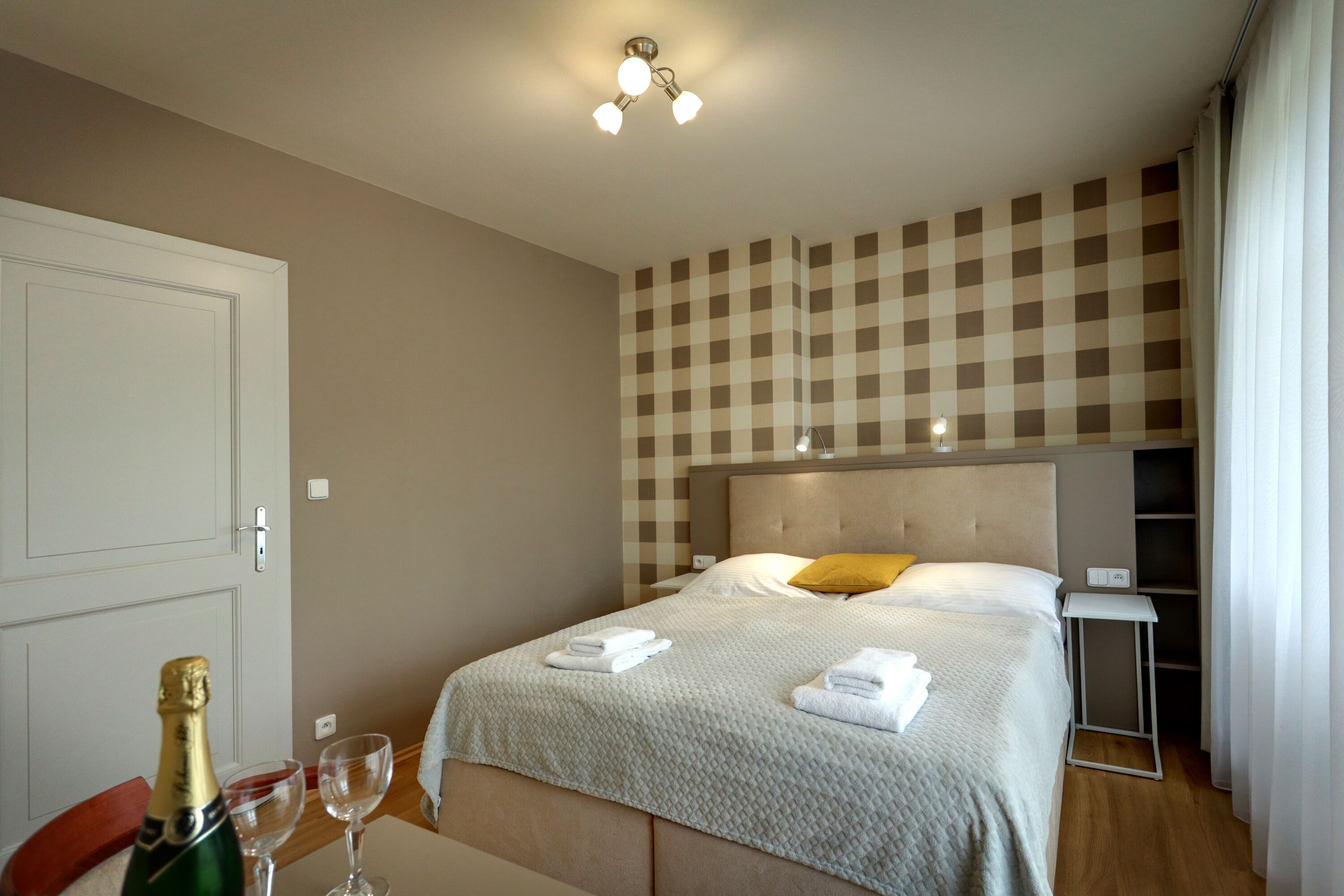 A hotel room with a checkered wall, a bed with a headboard, a bedside table, and a bottle of champagne.