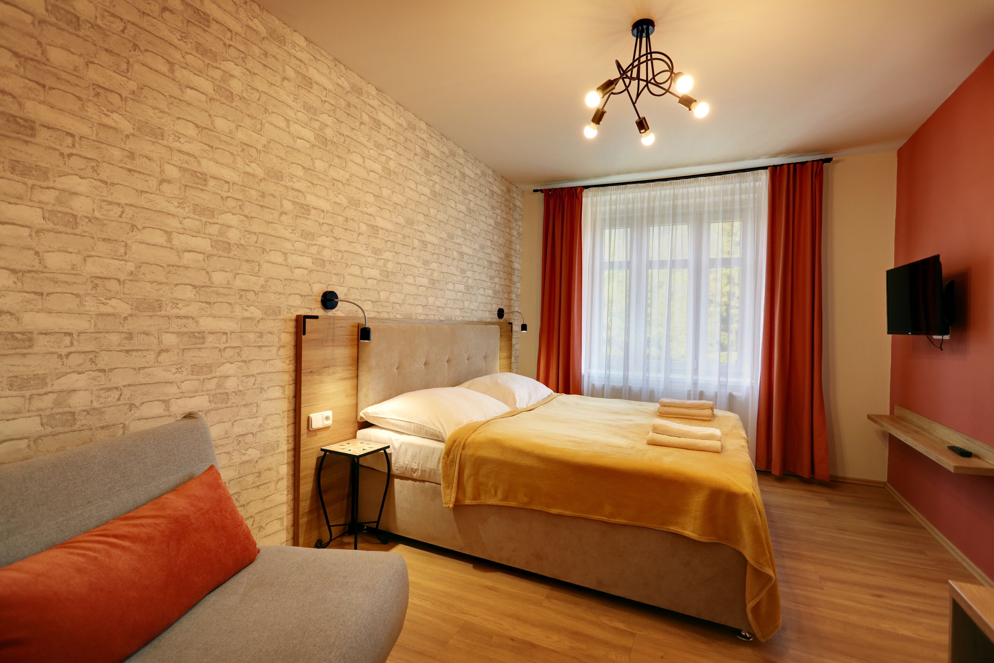 Ein Hotelzimmer mit Backsteinwand, einem Bett mit gelber Bettdecke, einem grauen Sofa, einem Nachttisch mit Lampe und einem an der Wand montierten Fernseher.