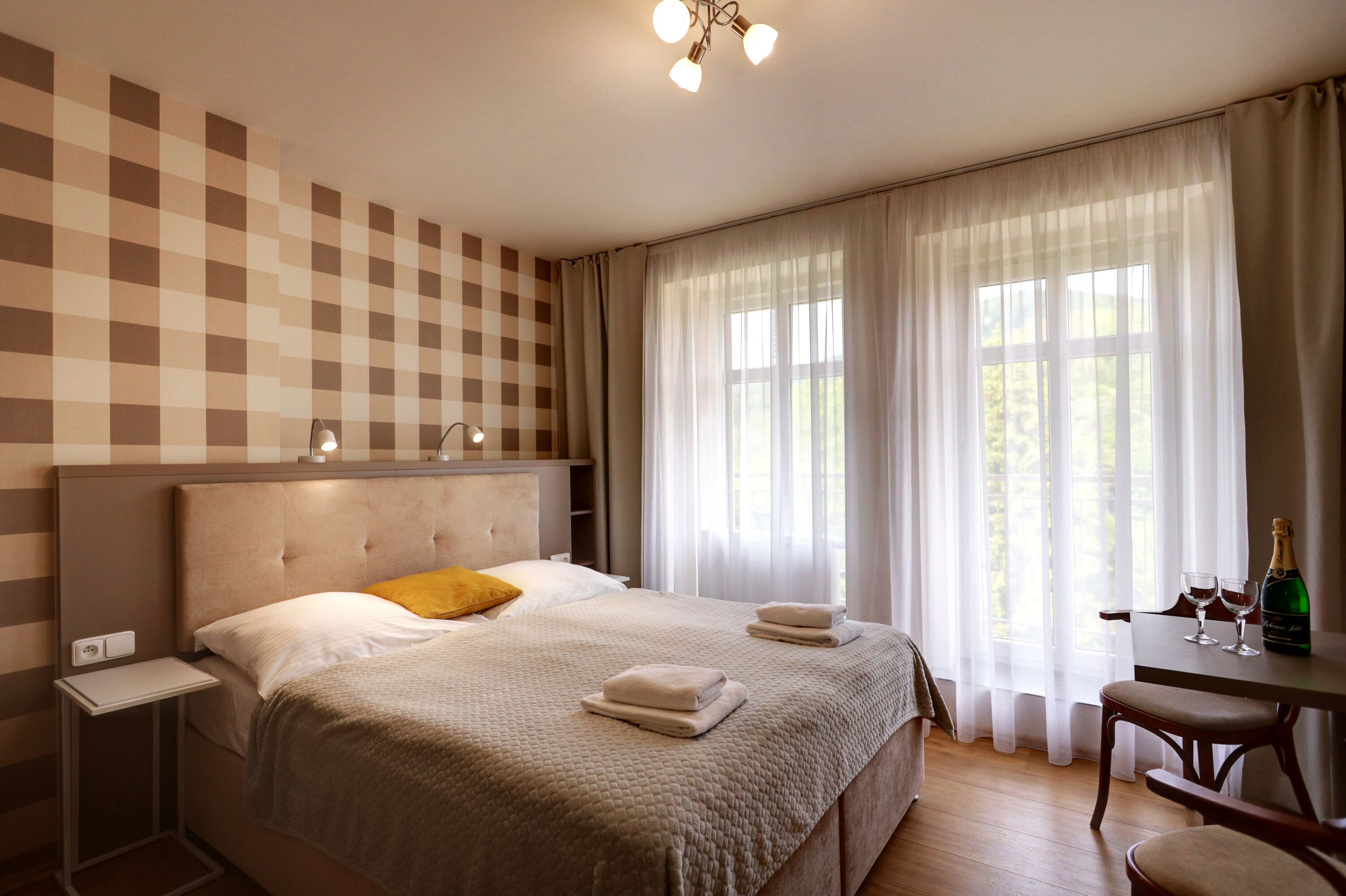 Une chambre d’hôtel avec un grand lit, une table de chevet, une chaise, une table avec une bouteille de champagne et des verres, et une fenêtre avec des rideaux.