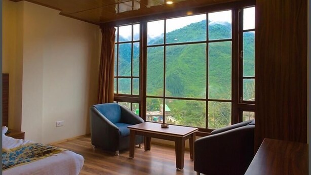 A room with a bed, a blue armchair, a small table, and a view of green hills through large windows.