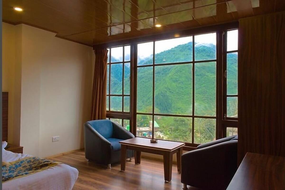 A room with a bed, a blue armchair, a small table, and a view of green hills through large windows.
