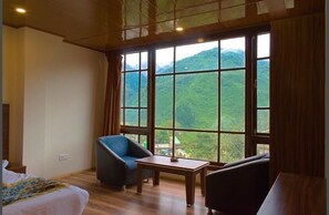 Une pièce comprenant un lit, un fauteuil bleu, une petite table et une vue sur des collines verdoyantes, visible à travers de grandes fenêtres.