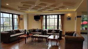 A spacious living area with multiple sofas and a coffee table, large windows with curtains, and a flat-screen TV mounted on the wall.