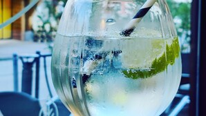 A glass of a clear beverage with ice and a straw, placed on a table with a view of a building and greenery outside.