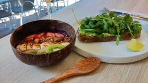 A dining table with a bowl of salad, a slice of toast with avocado and greens, a glass of orange juice, and a wooden spoon.
