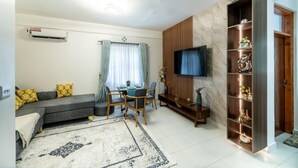 Ein Wohnzimmer mit einer Couch, einem Esstisch mit Stühlen, einem an der Wand befestigten Fernseher und einem Holzschrank mit Regalen.
