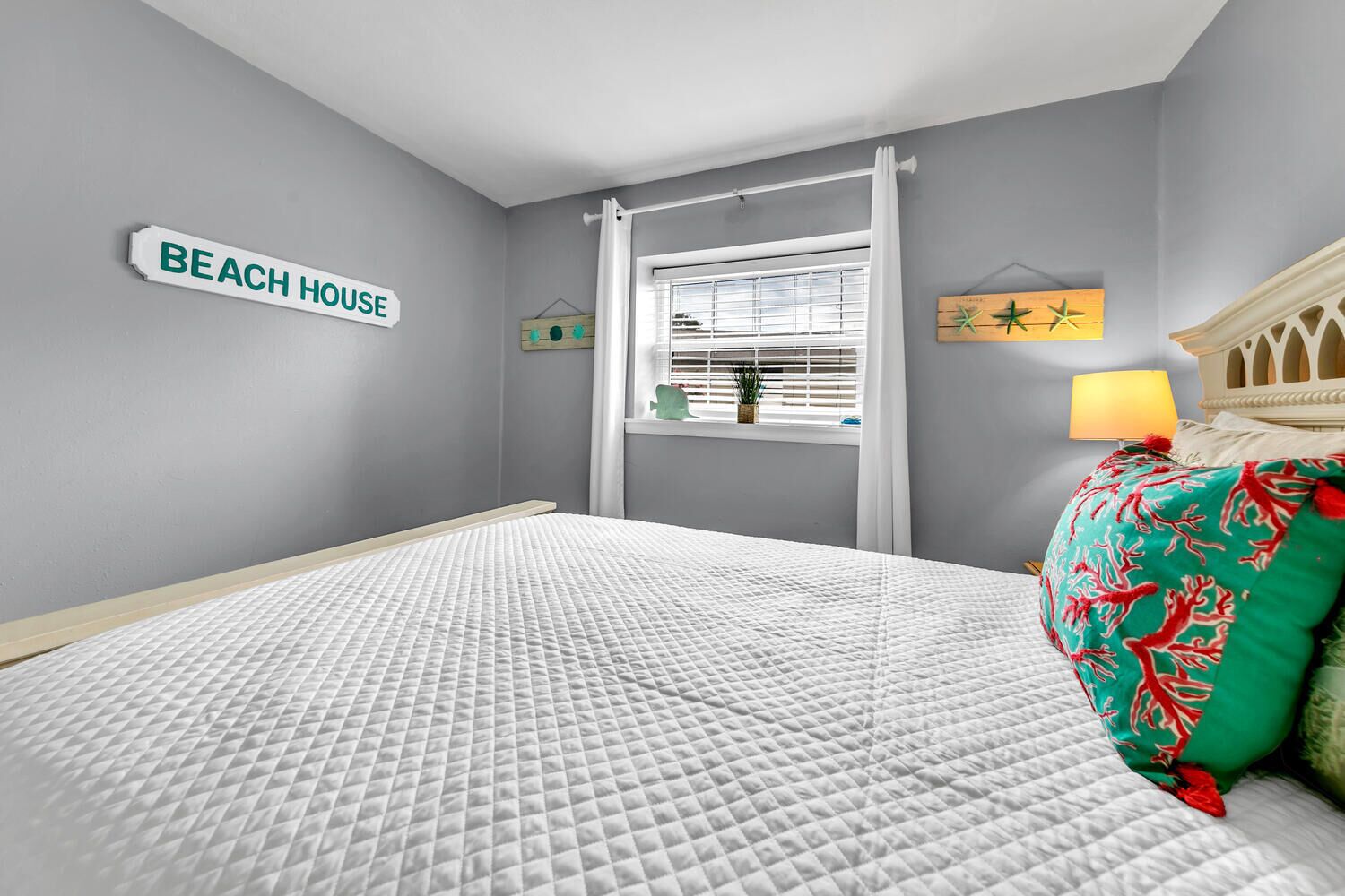 A bedroom with a bed, a window with blinds, a sign reading "BEACH HOUSE", and a lamp on the nightstand.