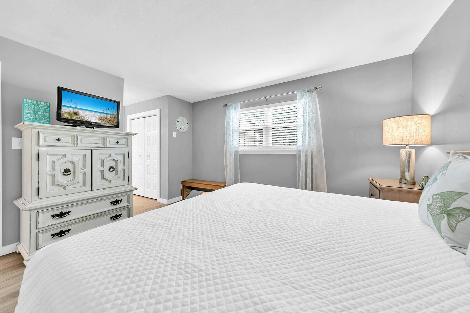 A bedroom with a large bed, a dresser, a nightstand, a lamp, a window with curtains, and a wall clock.