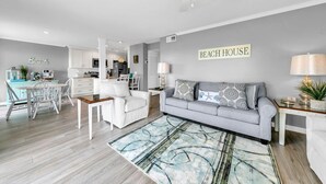 En moderne stue med en grå sofa, hvide lænestole og et spiseområde med bord og stole. Et køkken kan ses i baggrunden.