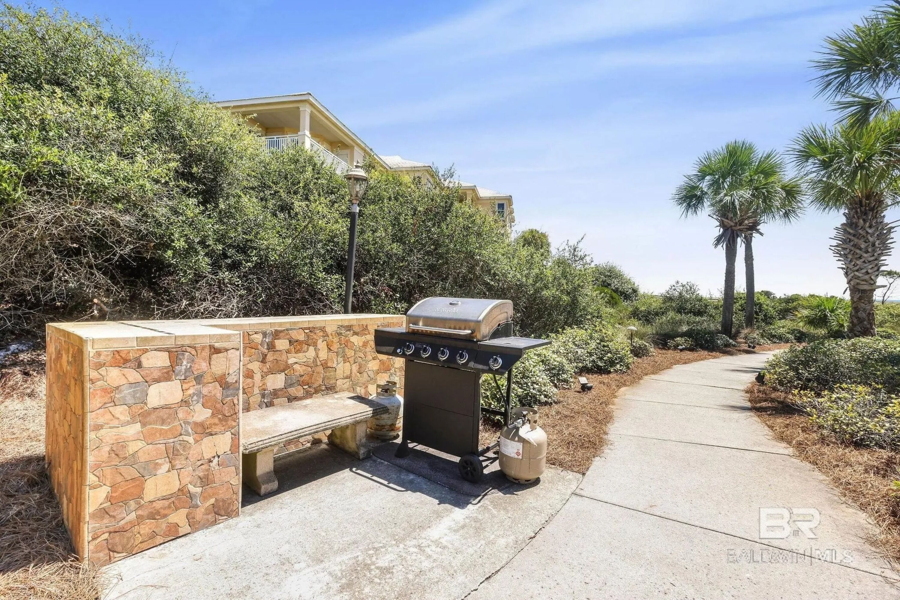An outdoor grill setup with a stone bench and a gas tank, surrounded by greenery and a pathway.