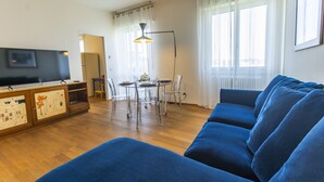 A living room with a blue sofa, a dining table with chairs, a TV on a cabinet, and a window with curtains.