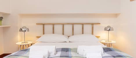 Ein ordentlich bezogenes Bett mit zwei Kissen und zwei gefalteten Handtüchern, das von je einer Nachttischlampe an beiden Seiten beleuchtet wird.