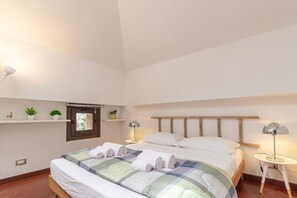 A neatly arranged bedroom with a bed, bedside tables, a window with blinds, and a small shelf with plants. - Il Camino Felice - 201 Autodromo F1 Villa Reale (Monza)