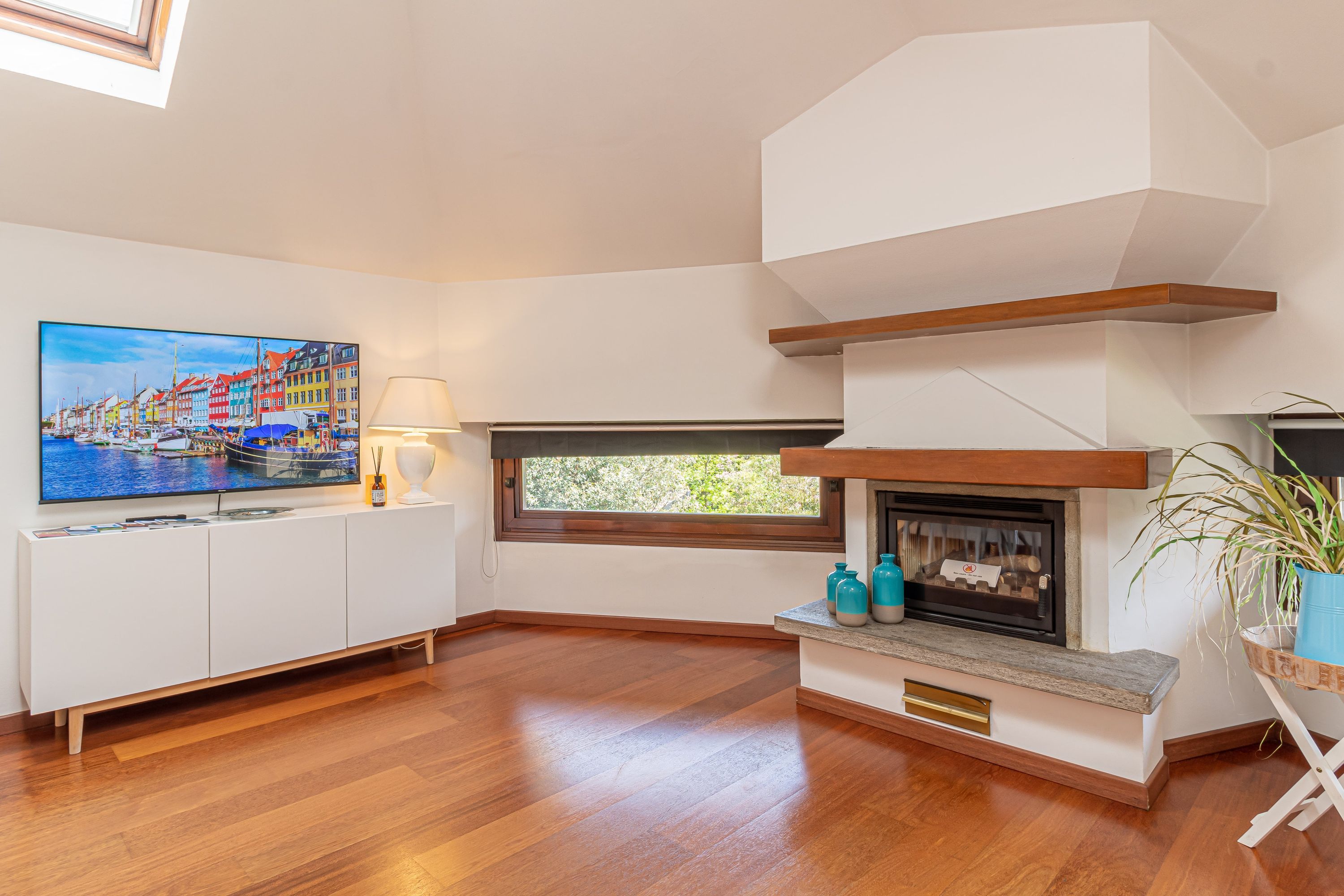 Sala de estar com lareira, televisão em um armário branco e janela com vista para o verde.