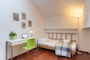 Une petite chambre à coucher, bien rangée, avec un lit simple, un bureau avec une chaise, une lampe de chevet et une étagère murale agrémentée d’une plante en pot.