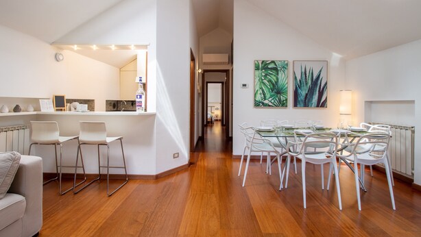 A modern kitchen with a central island, a dining area with a glass table and white chairs, and a hallway leading to another room. - Il Camino Felice - 201 Autodromo F1 Villa Reale (Monza)