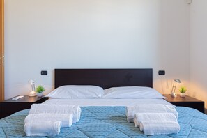A neatly made bed with white linens, a blue patterned bedspread, and four rolled towels on the bed. There are two bedside tables with lamps and small potted plants. - La Terrazza di Giotto - 501 (Lissone)