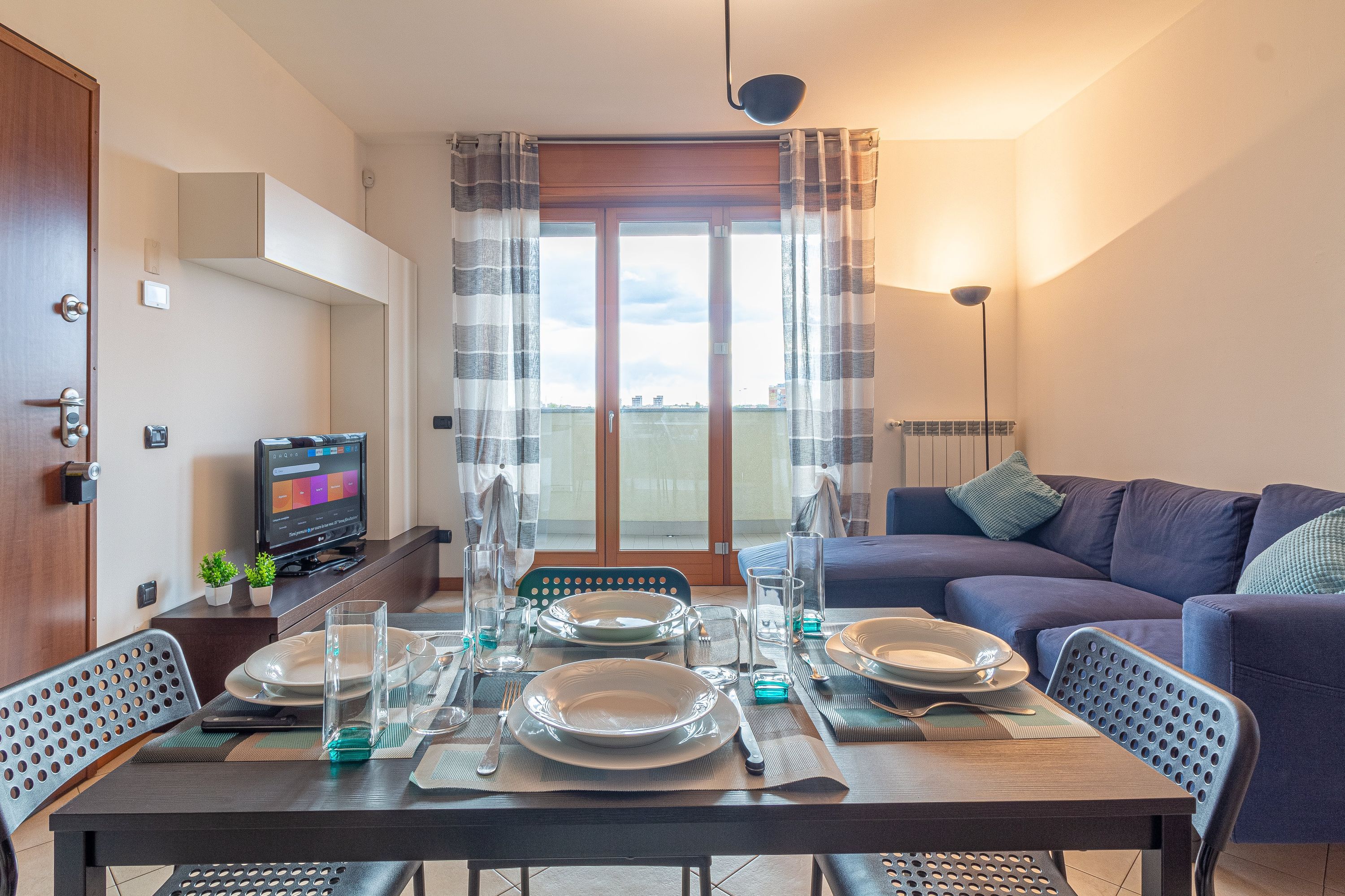A dining area with a table set for four, a sofa, a TV, and a balcony with curtains.