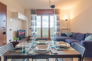 A dining area with a table set for four, a sofa, a TV, and a balcony with curtains. - La Terrazza di Giotto - 501 (Lissone)
