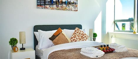 A neatly made bed with a patterned blanket, a nightstand with a lamp and a clock, a small plant, and a painting of a bridge.
