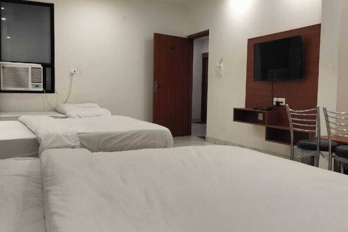 A hotel room with two single beds, a desk with chairs, a TV mounted on the wall, and an air conditioning unit.