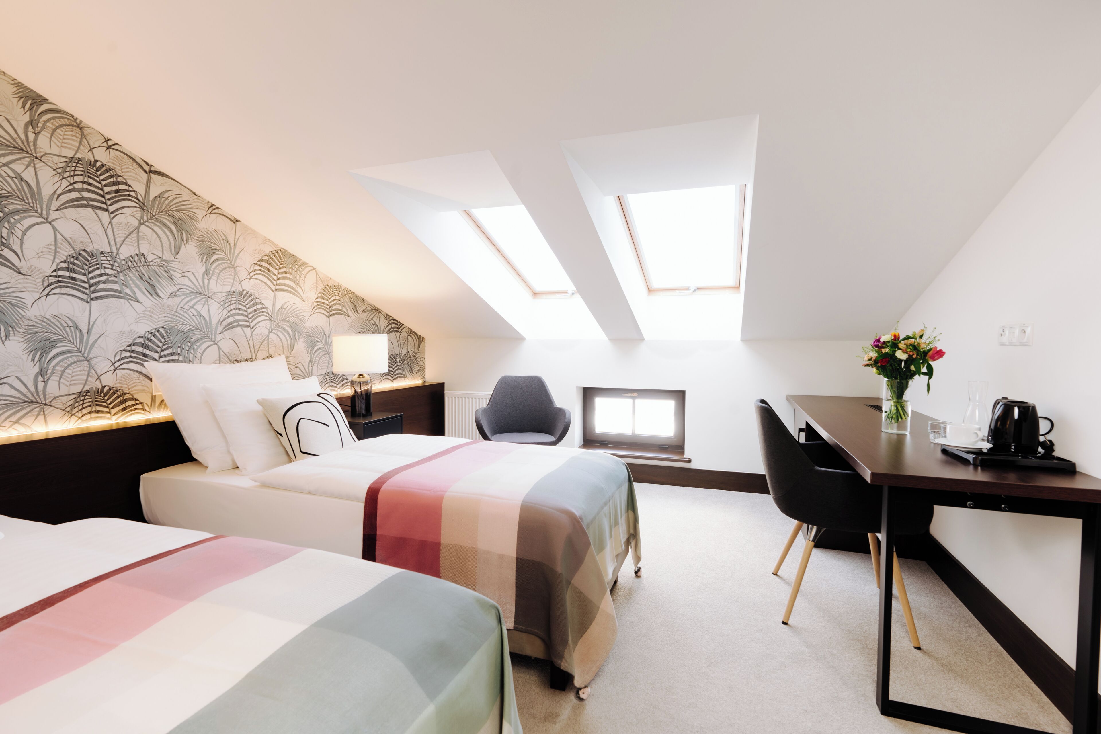 A modern hotel room with two beds, a desk, and a chair. There is a skylight above the desk and a large window.