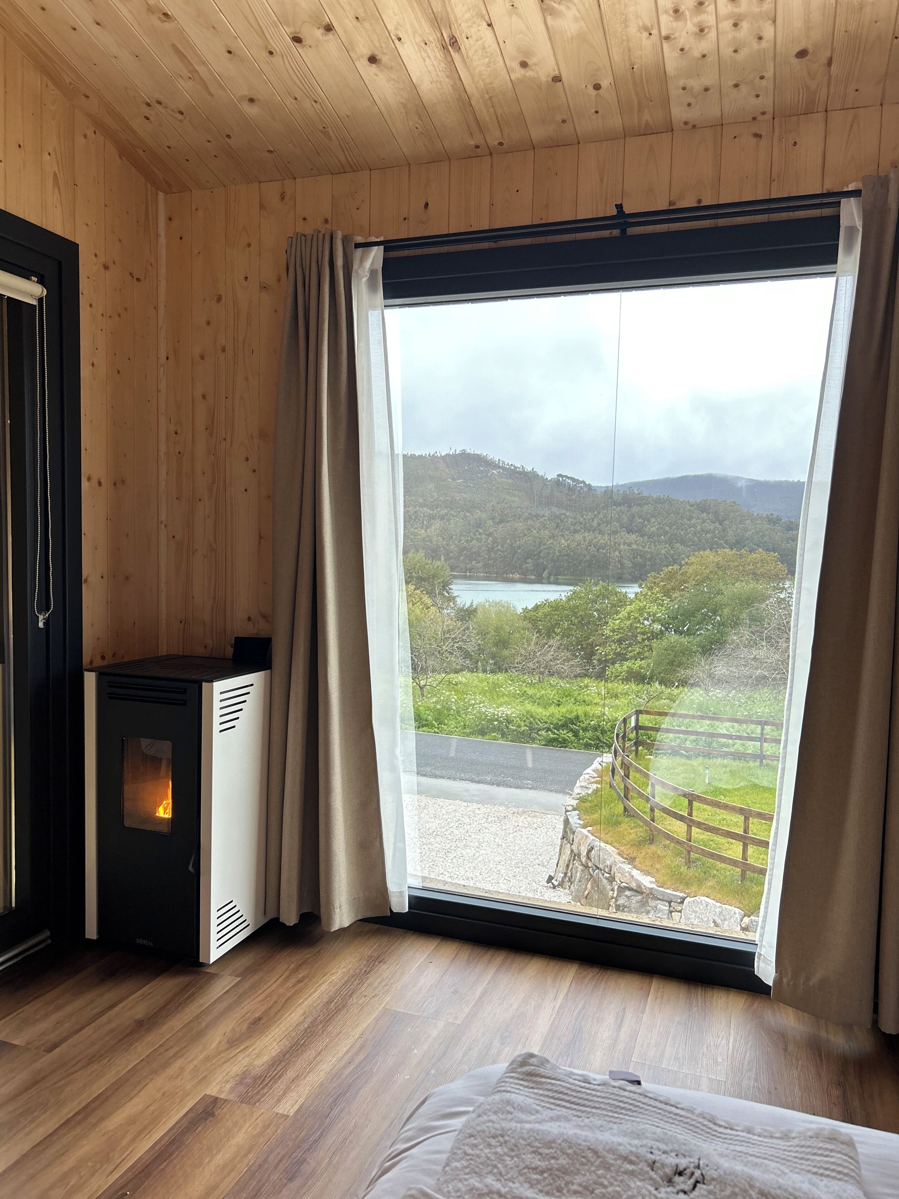 A room with wooden walls, a large window with a view of a lake and hills, a wood-burning stove, and a bed with a blanket.