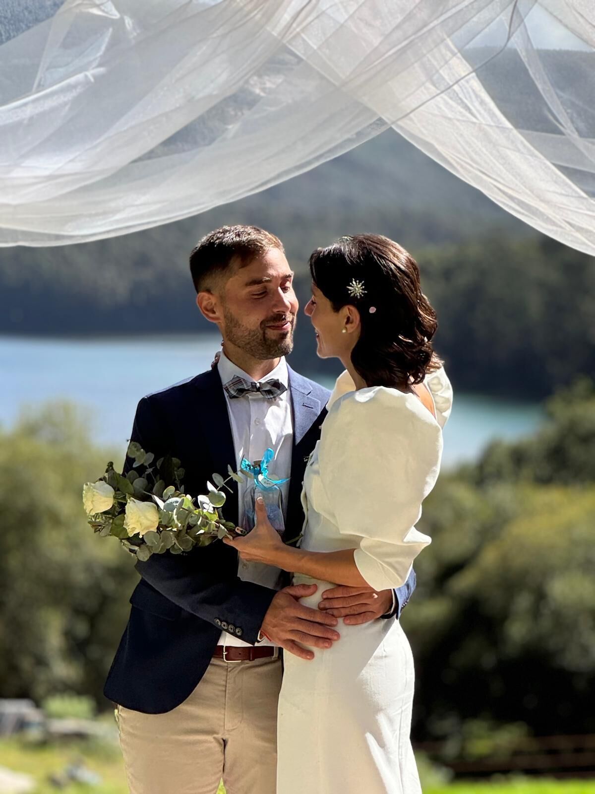 Una pareja se abraza bajo un dosel blanco translúcido, con un lago al fondo.