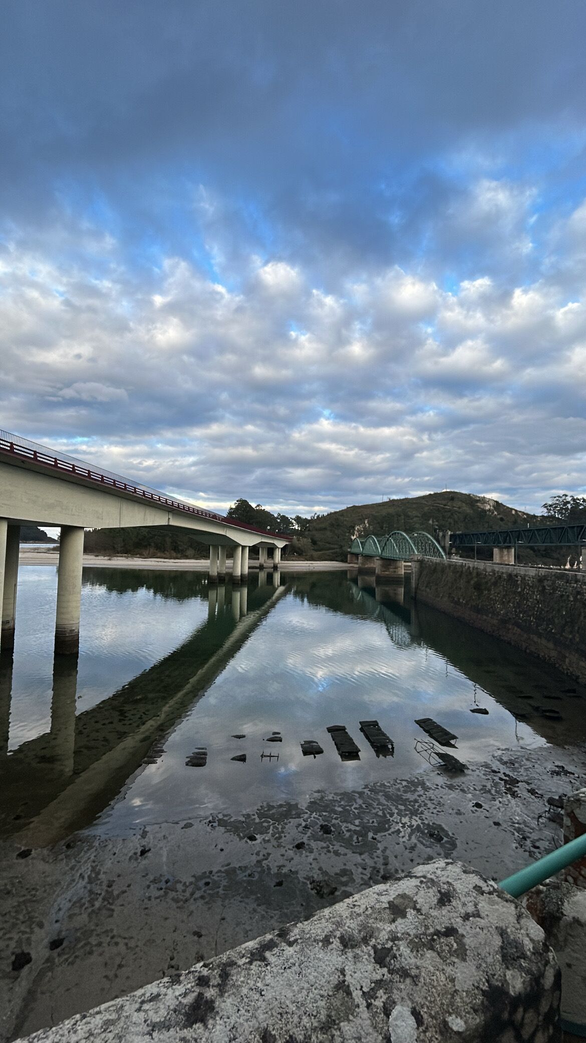 Un puente sobre un río tranquilo con un reflejo nítido, rodeado de colinas y un cielo nublado.