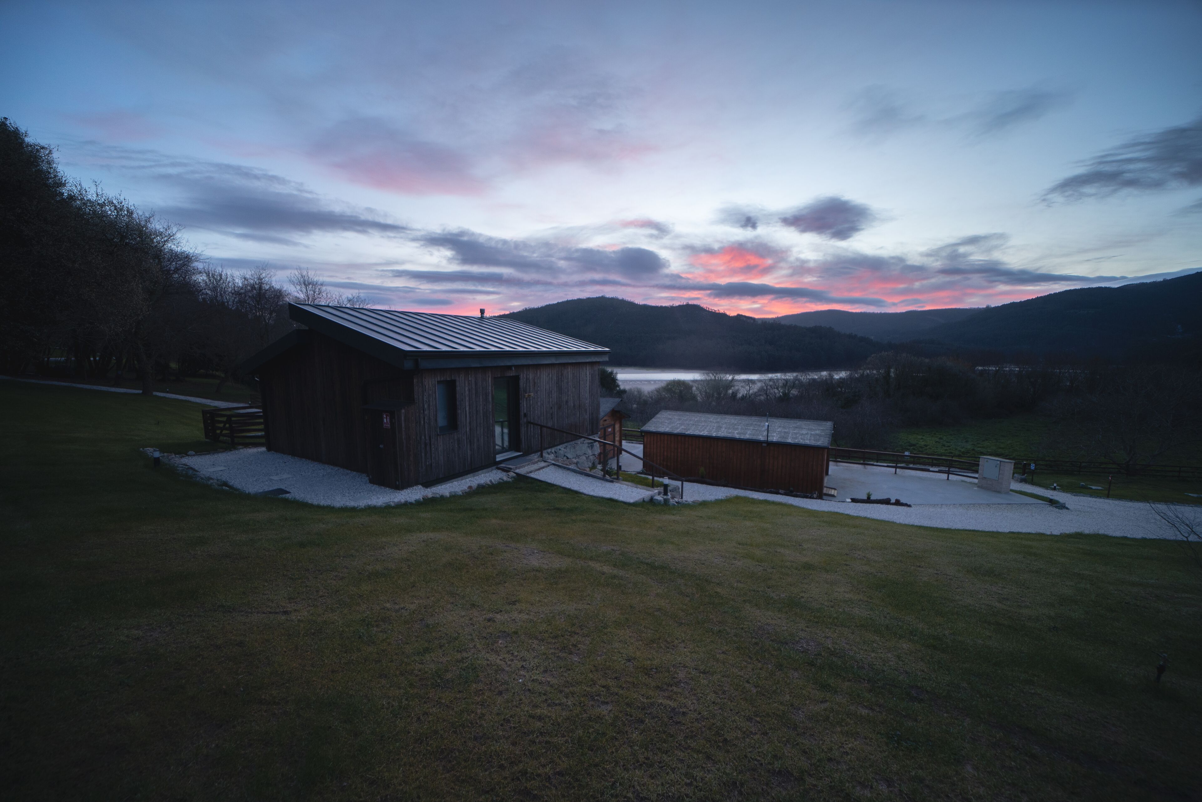 A rustic cabin with a metal roof, surrounded by a grassy area and a small wooden structure.