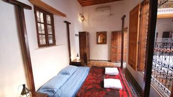Une chambre avec un lit, une table de chevet, une lampe fixée au mur et une fenêtre dotée d’une grille en métal.