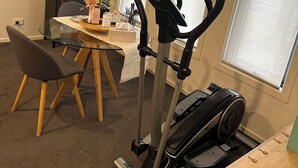 A home gym with an elliptical machine, a dining table with chairs, and a cabinet.