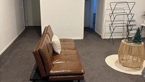 A room with a brown leather sofa, a small round side table with a plant, a white air conditioning unit, and a door leading to another room.