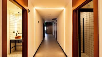 Un pasillo de hotel con diseño moderno, que presenta un largo pasillo con piso de baldosas, un área de lavamanos a la izquierda y un área de inodoros a la derecha.