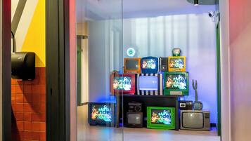 A hotel room entrance with a 'ROOMS' sign, a glass door, and a wall displaying various retro televisions.