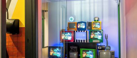 A hotel room entrance with a 'ROOMS' sign, a glass door, and a wall displaying various retro televisions.