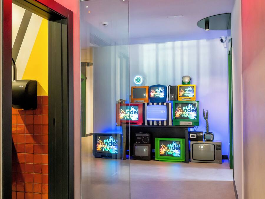 A hotel room entrance with a 'ROOMS' sign, a glass door, and a wall displaying various retro televisions.