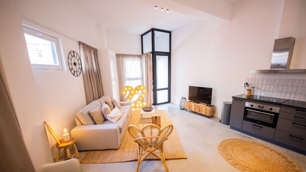 A modern living room with a sofa, coffee table, and wicker chairs. There is a kitchen area with a stove and cabinets.