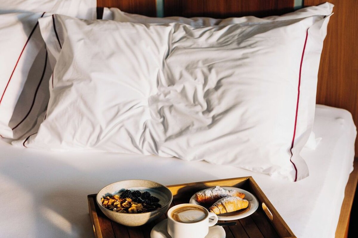 Een net opgemaakt bed met witte lakens en kussens, een dienblad met ontbijtartikelen, waaronder croissants en koffie, en een schaal met bessen.