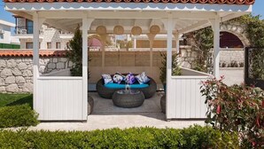 A covered outdoor seating area with a white pergola, stone walls, and a view of the garden hotel in the background. - Mara Bahçe Alaçatı (Cesme)