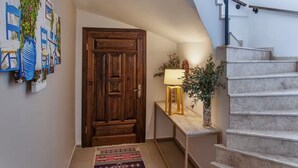 A hallway with a wooden door, a small table with a lamp, a vase with plants, and a staircase. - Mara Bahçe Alaçatı (Cesme)